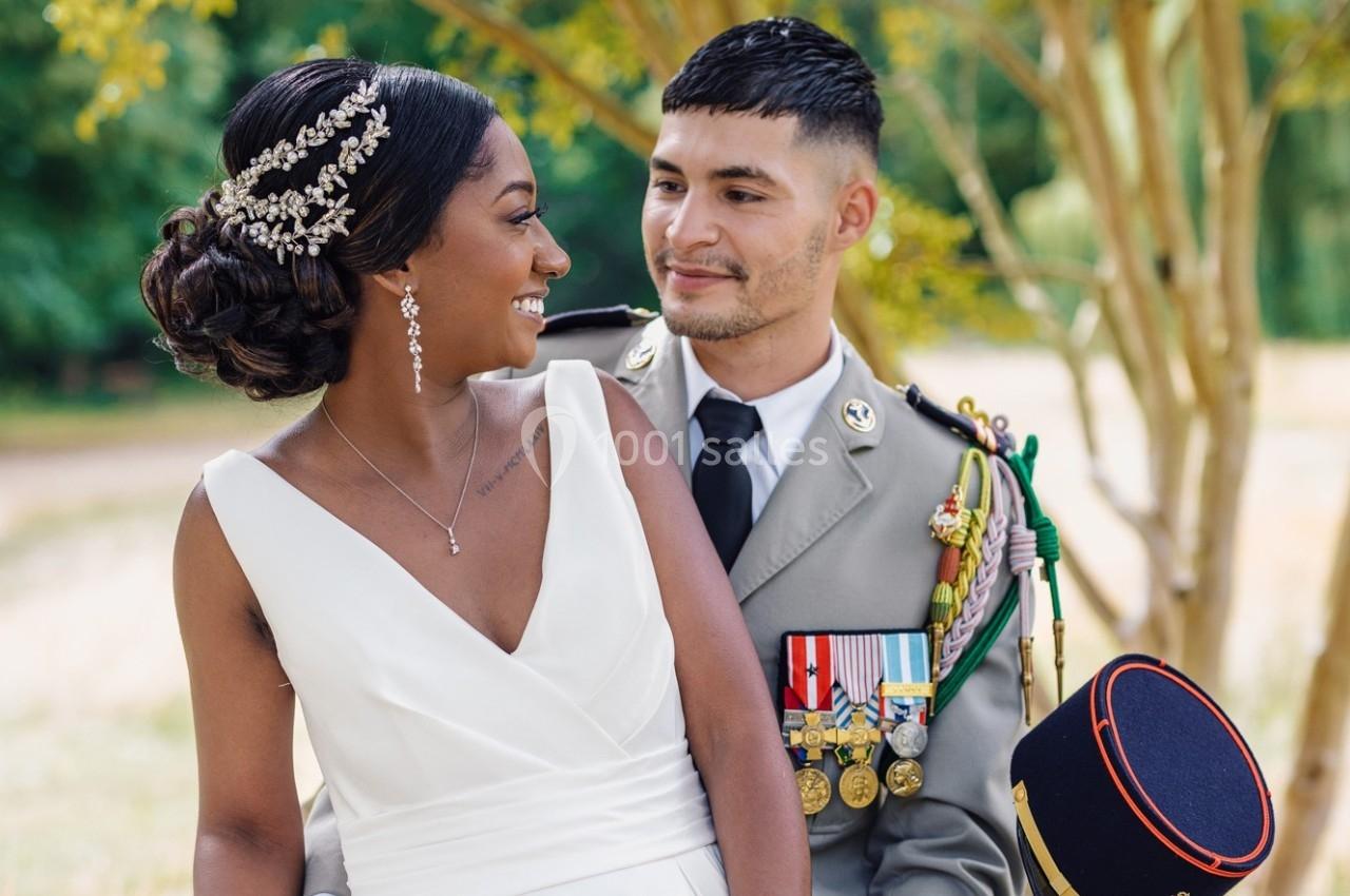 Une mariée en robe blanche et un militaire en uniforme posent ensemble dans un cadre extérieur verdoyant.