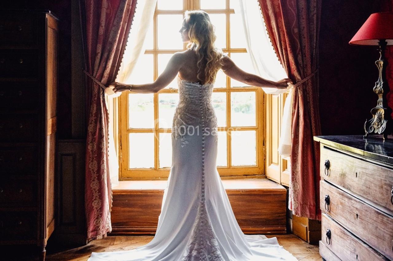 Femme en robe de mariée blanche debout devant une fenêtre ouverte, encadrée par des rideaux rouges dans une pièce sombre.