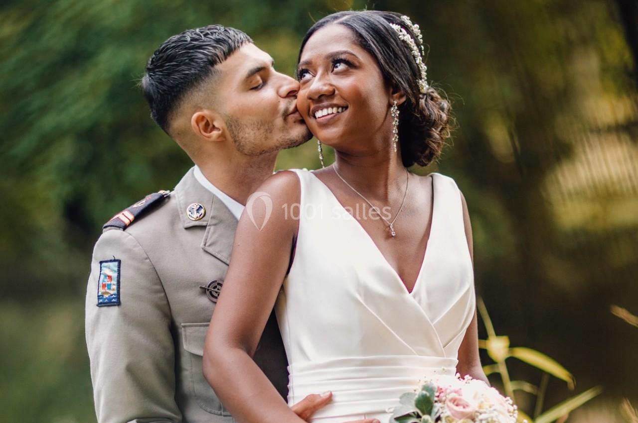 Un couple en tenue de mariage, l'homme en uniforme embrasse tendrement la femme souriante dans un cadre naturel.