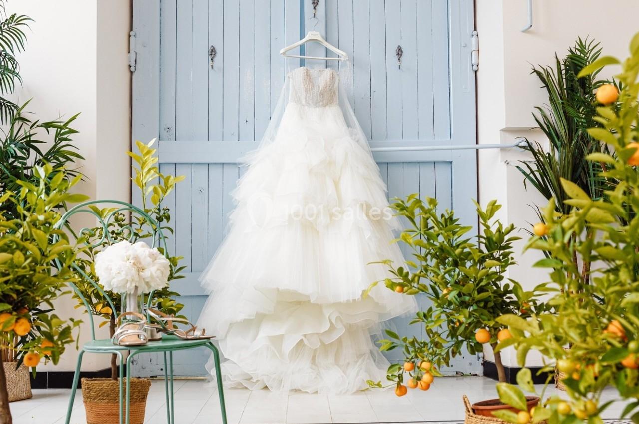 Robe de mariée blanche à volants suspendue devant une porte bleue, entourée de plantes et d'orangers en pot.