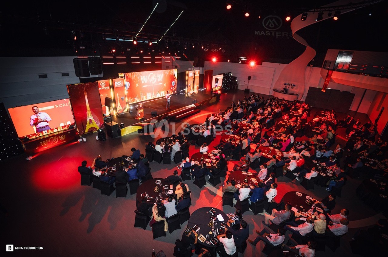 Salle de conférence illuminée en rouge avec un public assis à des tables rondes, regardant une scène avec un orateur.