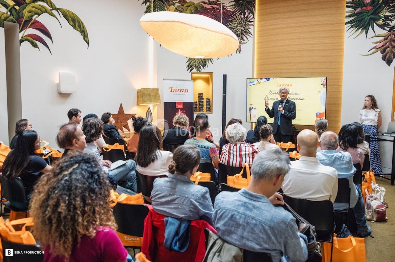 Un conférencier s'adresse à un public assis dans une salle décorée de motifs tropicaux.