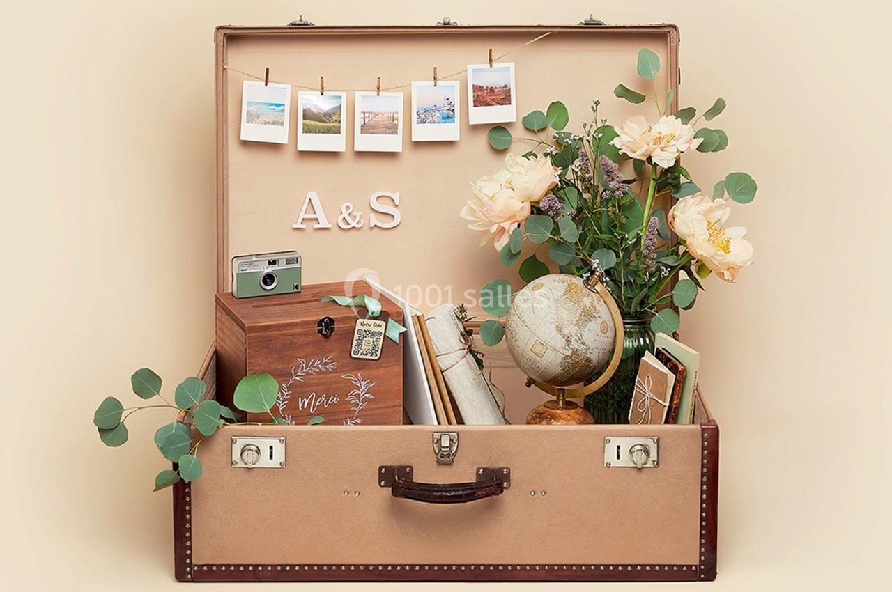 Valise ouverte contenant des objets décoratifs : globe terrestre, appareil photo vintage, fleurs et photos suspendues.