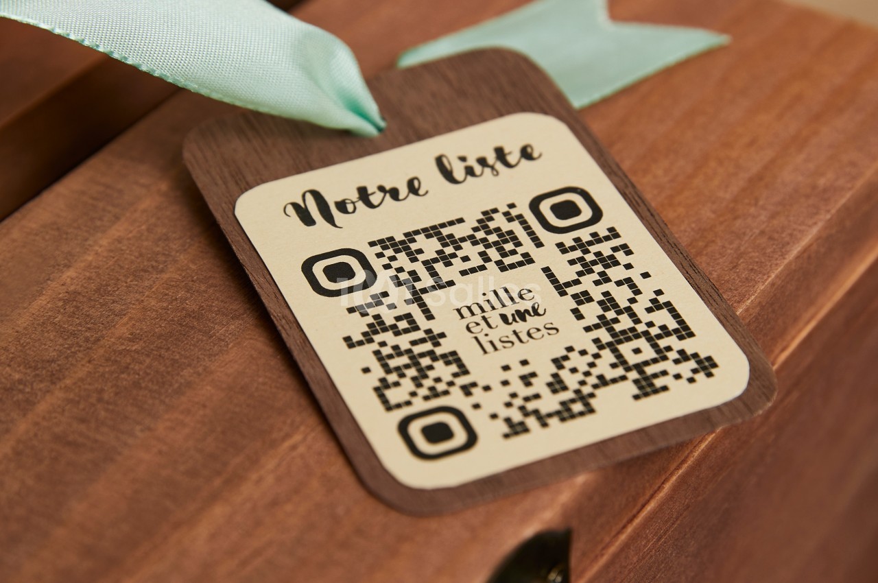 Étiquette en bois avec un QR code et l'inscription ’Notre liste’, attachée à un ruban vert sur une surface en bois.