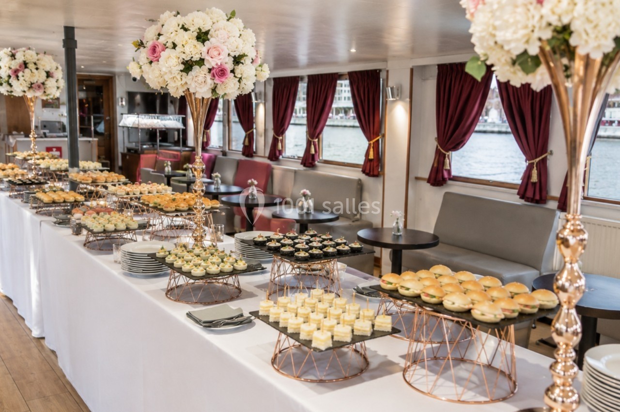 Buffet élégant avec desserts et amuse-bouches disposés sur une table dans une salle décorée de fleurs et de rideaux rouges.