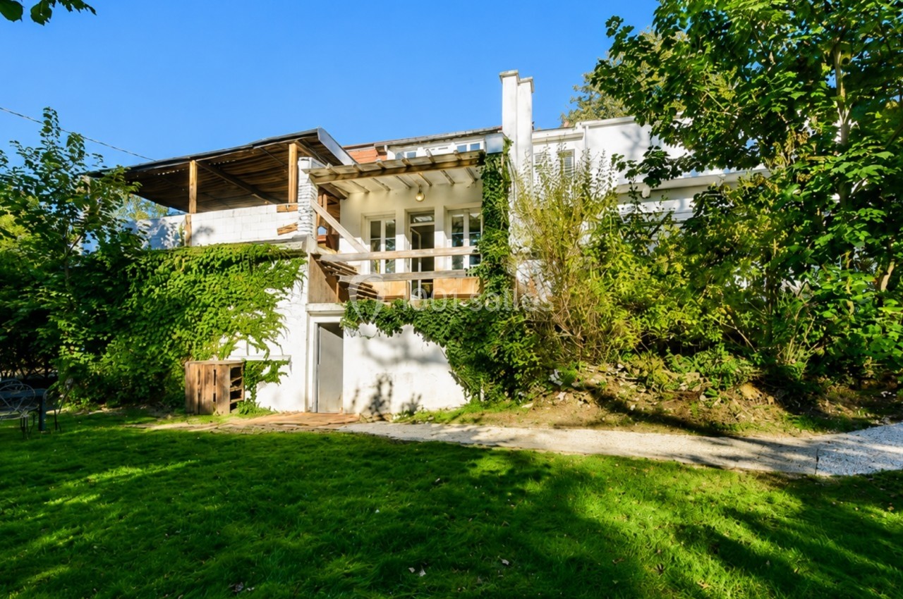 Façade d'une maison blanche entourée de verdure, avec terrasse en bois et jardin ensoleillé.