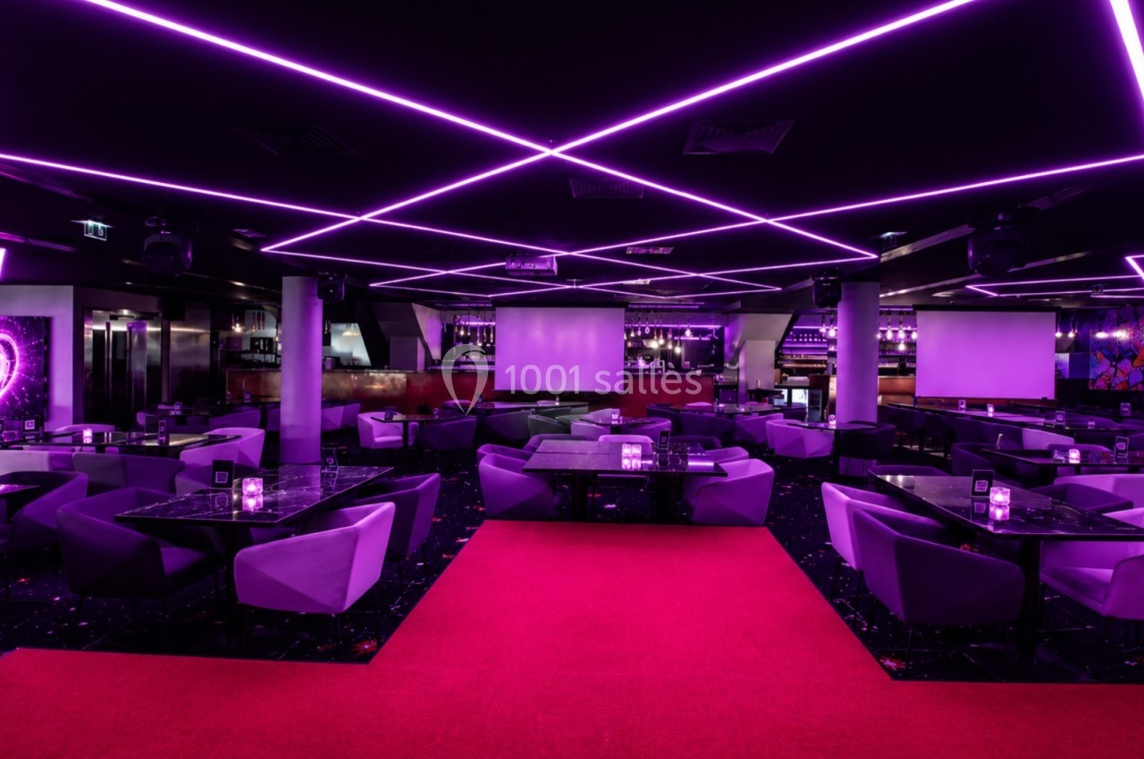 Salle moderne avec éclairage néon violet, tables et fauteuils disposés autour d'une scène et d'un bar en arrière-plan.