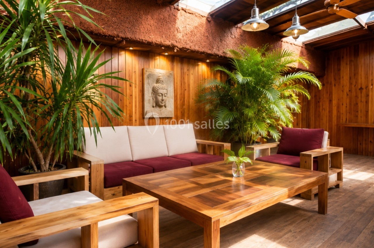Espace salon chaleureux avec mobilier en bois, coussins rouges, plantes vertes et lumière naturelle entrant par le toit.