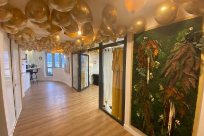 Salon lumineux avec mobilier en bois, plantes vertes, canapés, table basse et cuisine ouverte en arrière-plan.