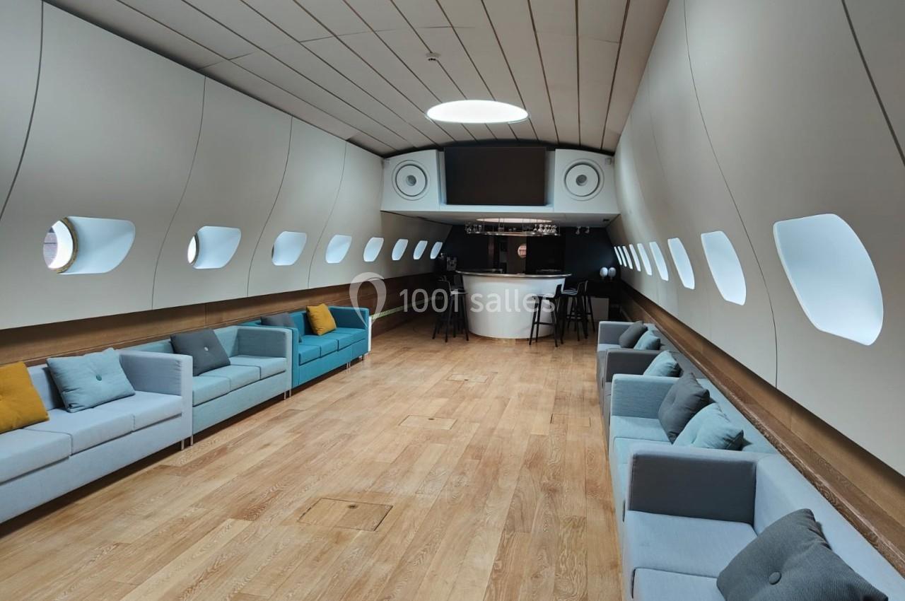 Intérieur spacieux d'une pièce en forme de cabine d'avion avec des canapés, un bar central et un éclairage doux.