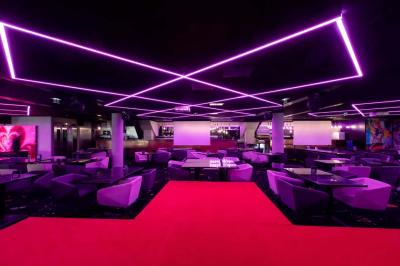 Miniature Le Grand Club du 16ème Salon lumineux avec mobilier en bois, plantes vertes, canapés, table basse et cuisine ouverte en arrière-plan.