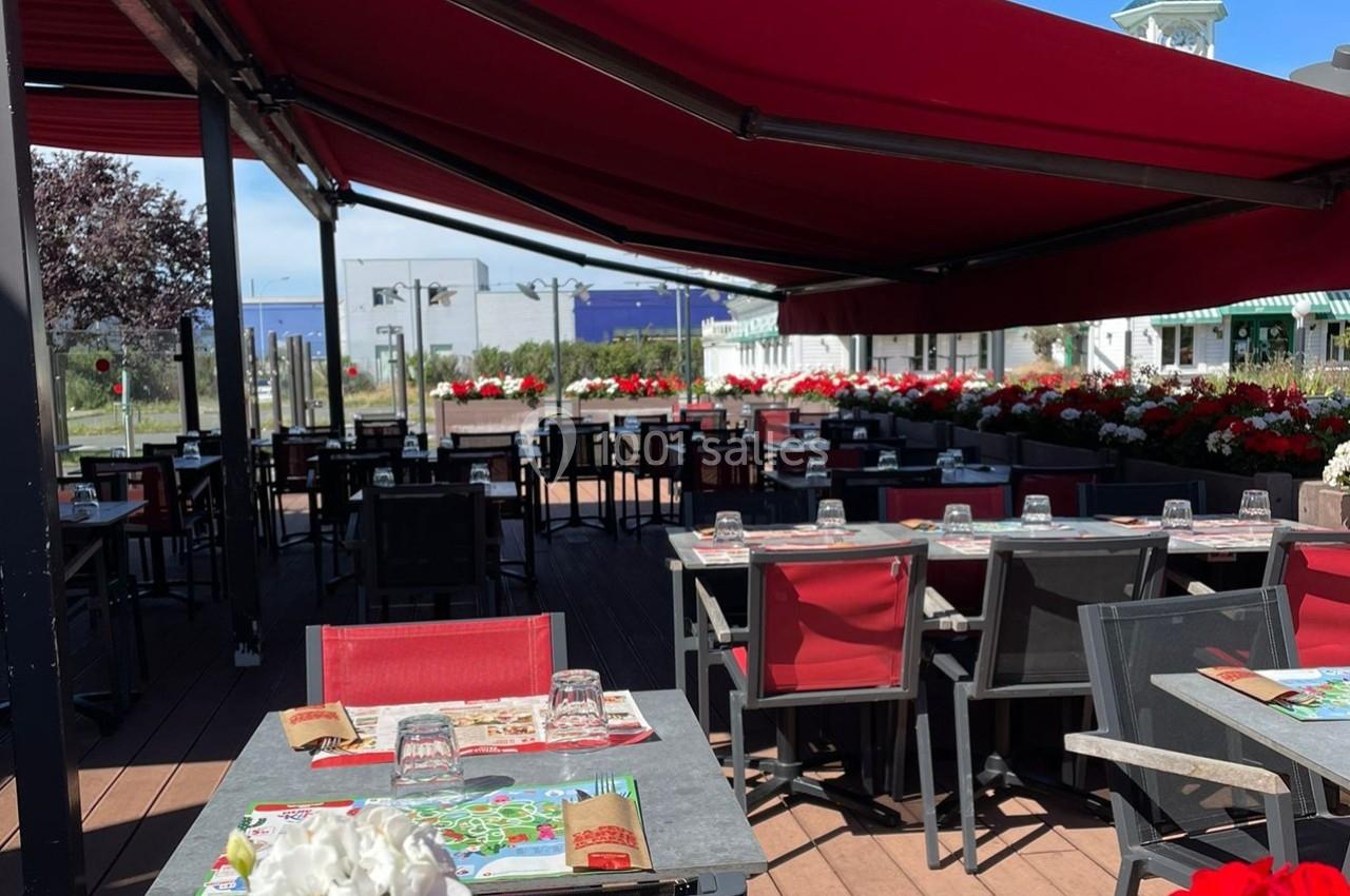 Terrasse extérieure d'un restaurant avec tables dressées, chaises rouges et noires, sous un grand auvent rouge.