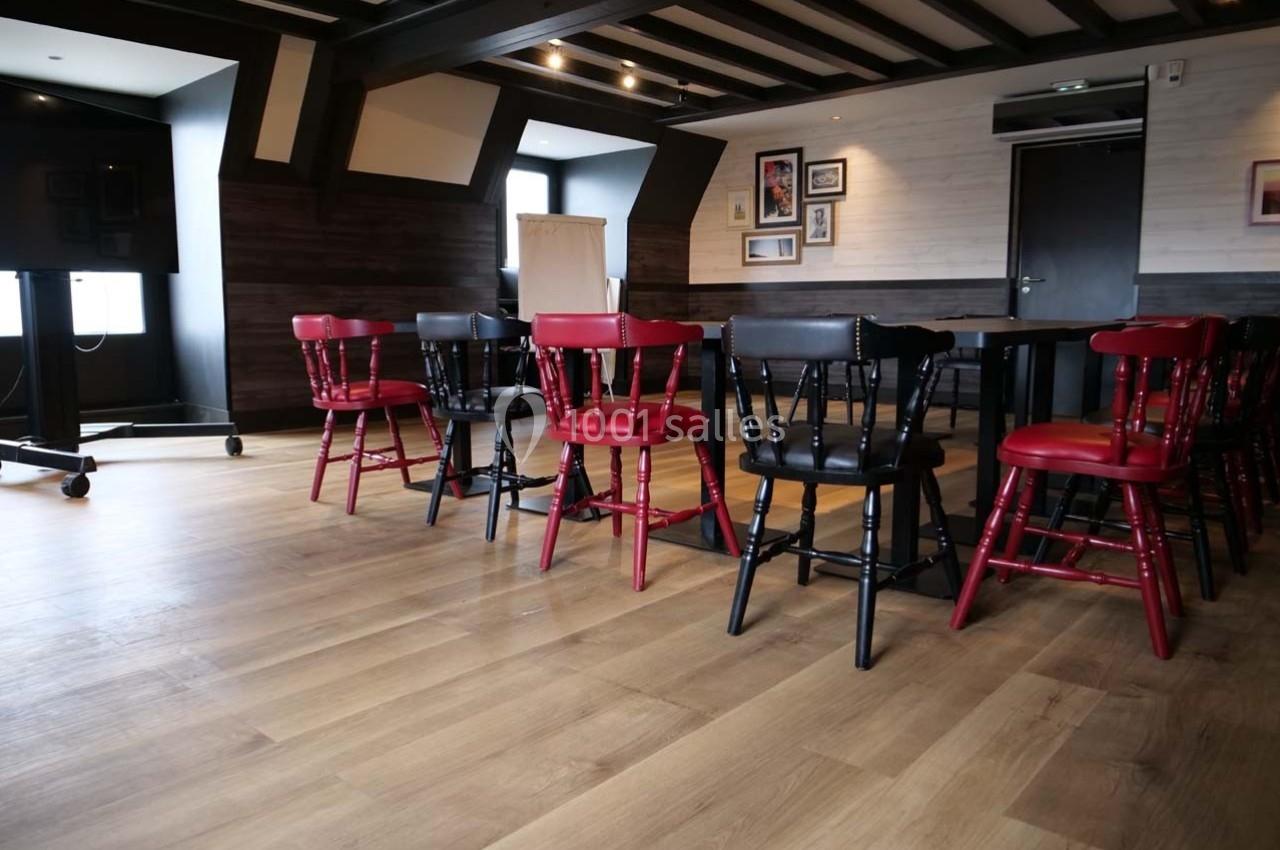 Salle de réunion avec chaises rouges et noires autour d'une table, parquet en bois clair et éclairage tamisé.