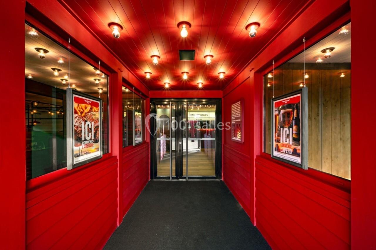 Entrée d'un bâtiment avec murs rouges, affiches publicitaires et portes vitrées au fond, éclairée par des lampes au plafond.