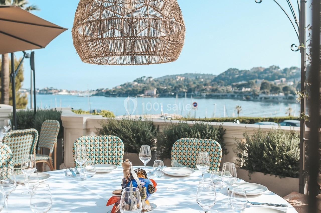Table dressée en extérieur avec vue sur une baie, des collines et des voiliers, sous une suspension en osier.