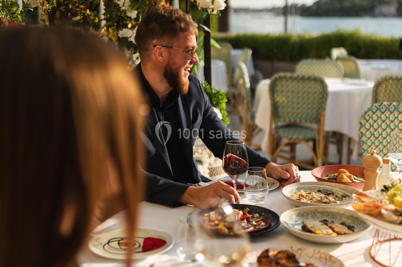 Un homme souriant partage un repas en terrasse, entouré de plats variés et d'une vue sur un paysage ensoleillé.