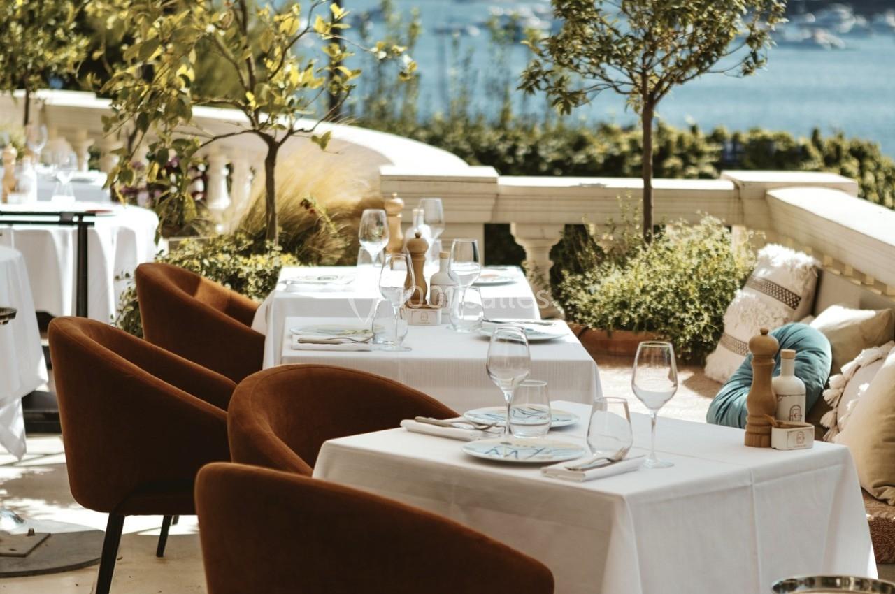 Terrasse de restaurant élégante avec tables dressées, chaises confortables et vue sur un paysage côtier en arrière-plan.