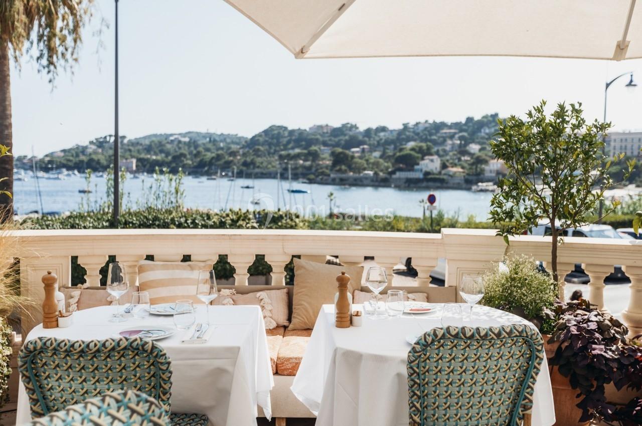 Terrasse ensoleillée d'un restaurant avec vue sur une baie, des collines verdoyantes et des voiliers au loin.