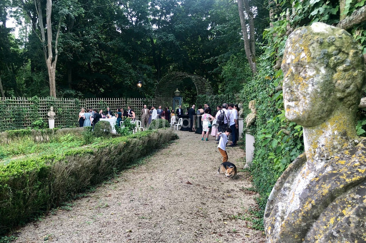 Un jardin verdoyant avec une statue en premier plan et un rassemblement de personnes au fond près d'une arche.