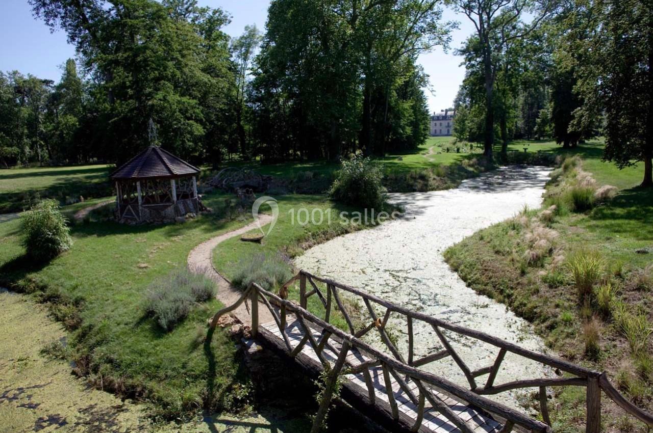Un pont en bois traverse un ruisseau bordé de végétation dans un parc avec un kiosque et un bâtiment en arrière-plan.