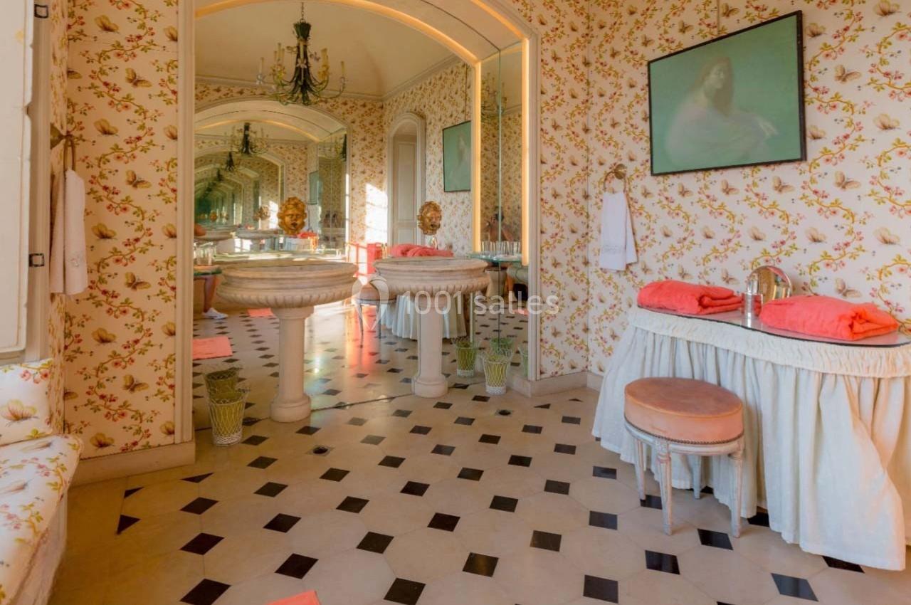 Salle de bain rétro avec papier peint fleuri, miroirs, lavabos sur pied, tabouret et serviettes orange pliées.