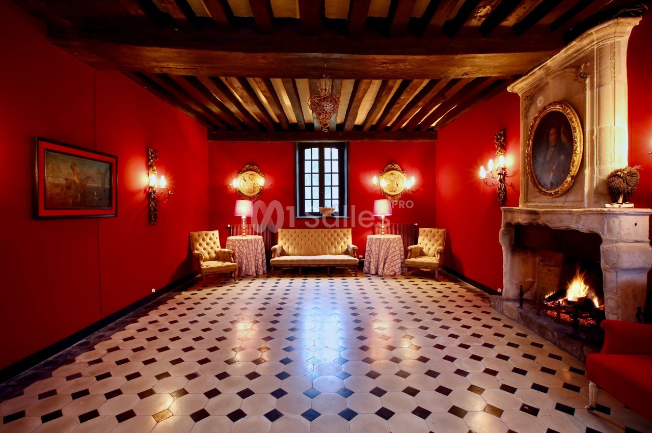 Salle élégante avec murs rouges, cheminée allumée, mobilier classique et plafond à poutres apparentes.