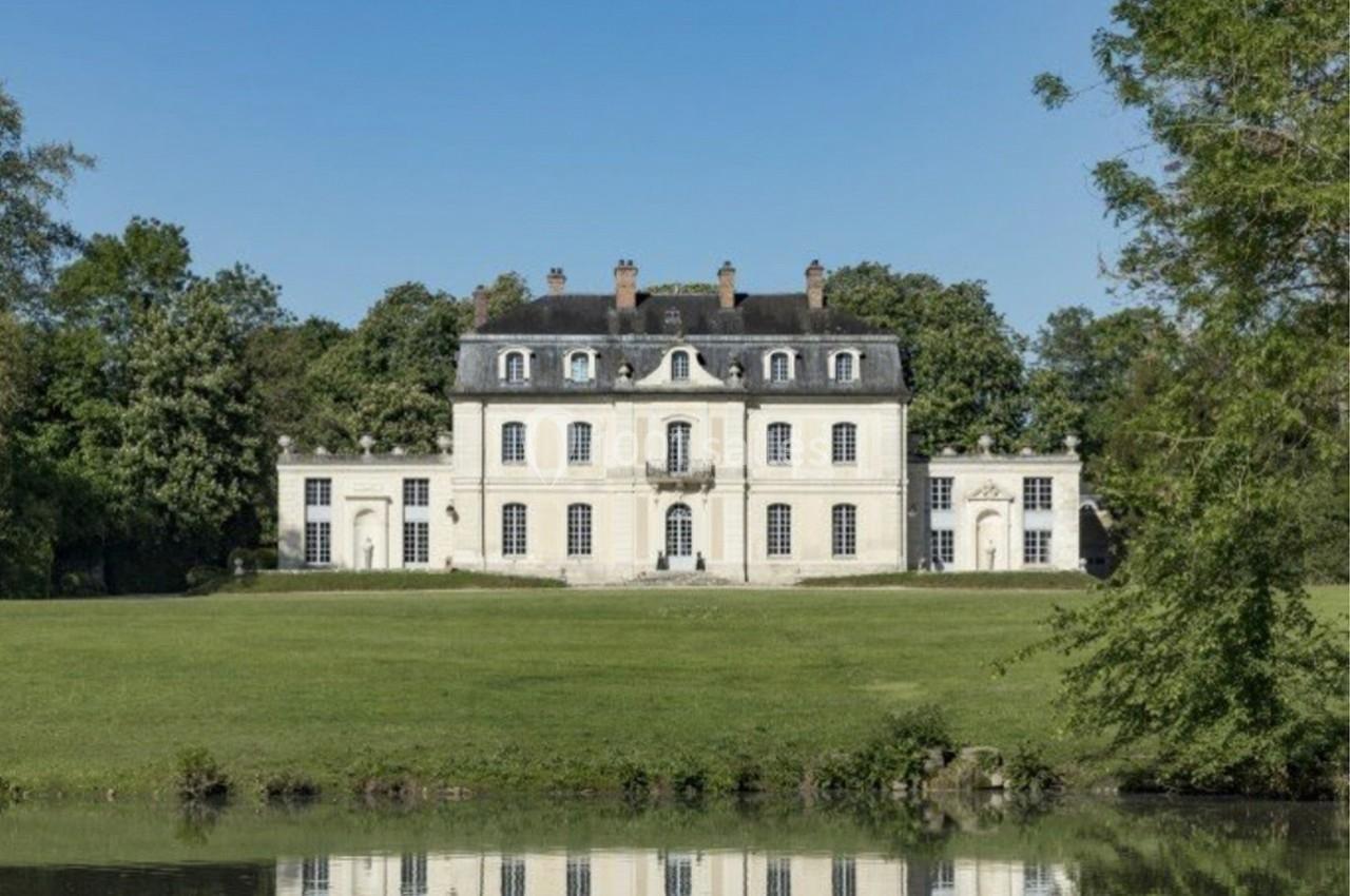Façade d'un château blanc entouré de verdure, avec un étang au premier plan sous un ciel dégagé.