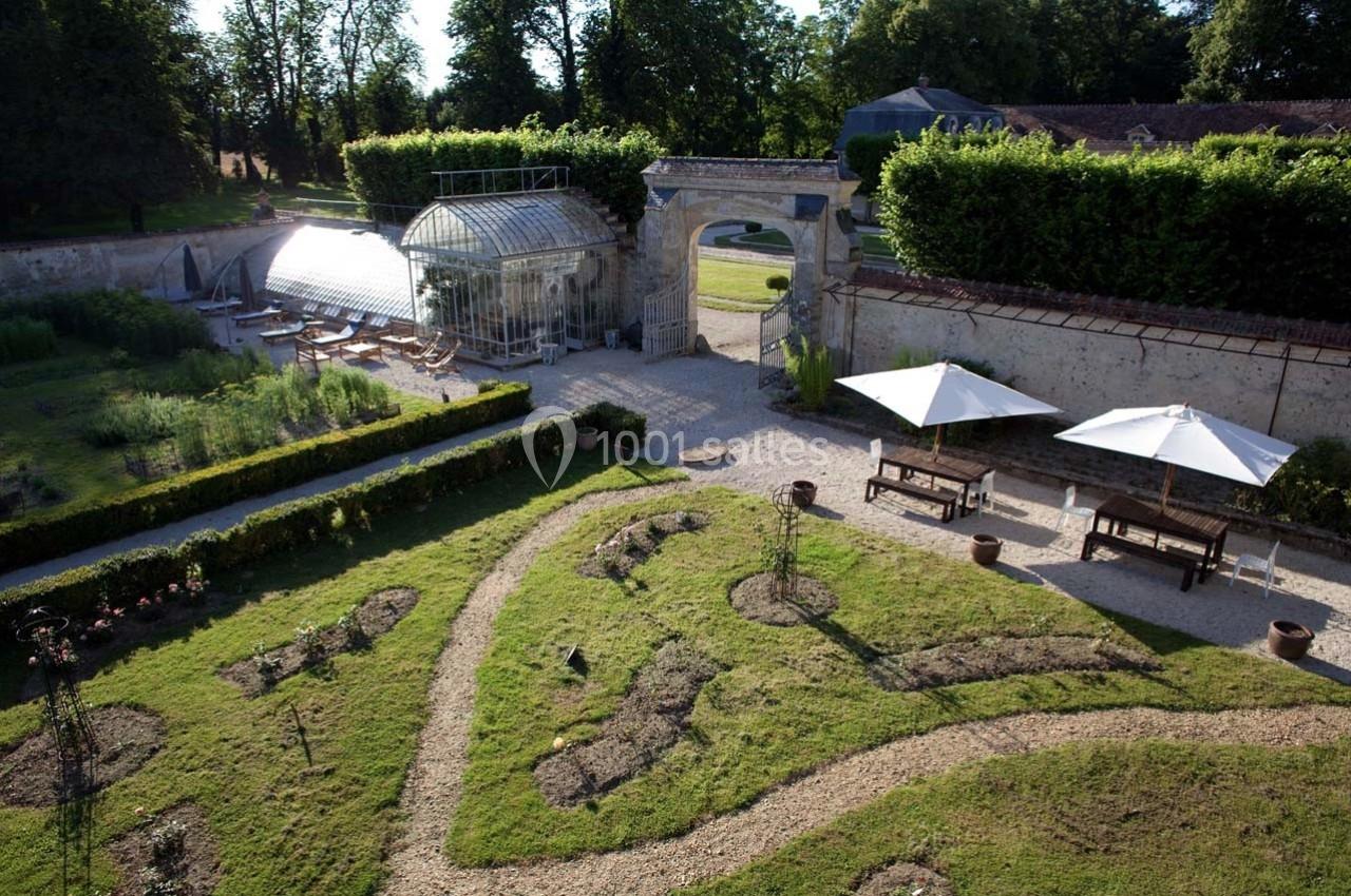 Jardin aménagé avec allées sinueuses, parterres de fleurs, serre en verre et mobilier extérieur sous parasols.