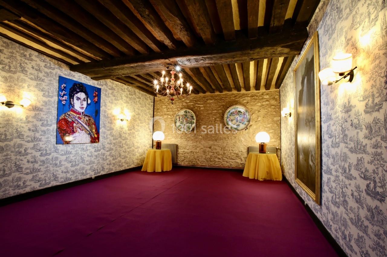 Salle décorée avec des murs en pierre et papier peint, lustre, tableaux, et tables rondes avec lampes.