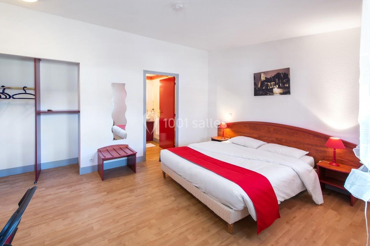 Chambre lumineuse avec lit double, linge blanc et rouge, parquet, armoire ouverte, et accès à une salle de bain.