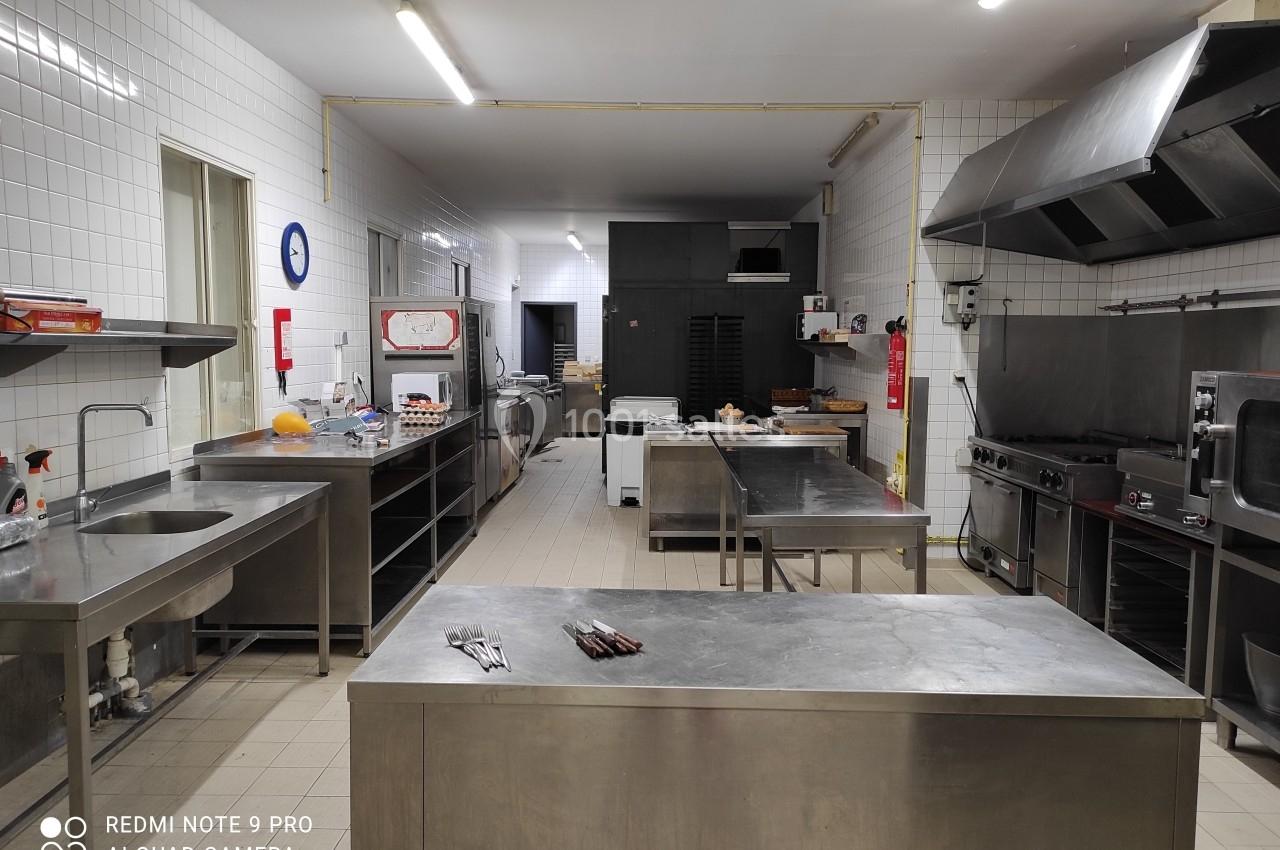 Cuisine professionnelle équipée avec plans de travail en inox, fours, plaques de cuisson et étagères de rangement.