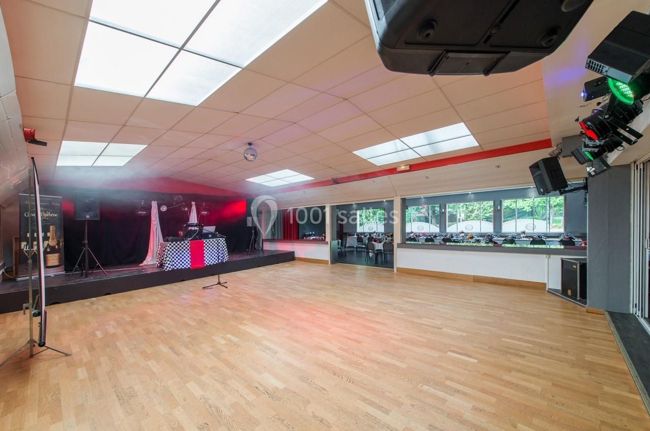Salle spacieuse avec parquet, scène équipée pour événements et lumière naturelle provenant de grandes fenêtres.