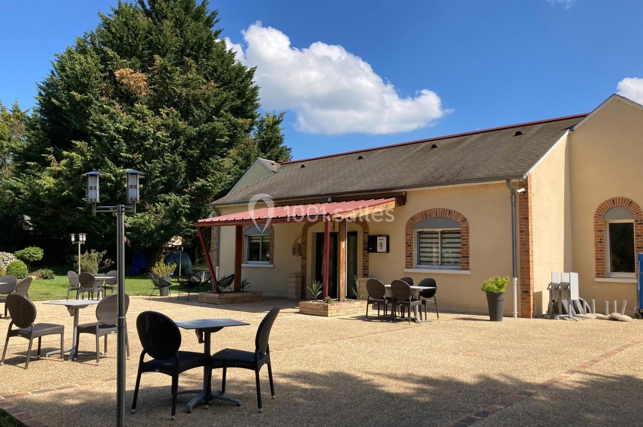Terrasse avec tables et chaises devant un bâtiment beige à toit en ardoise, entouré de végétation.
