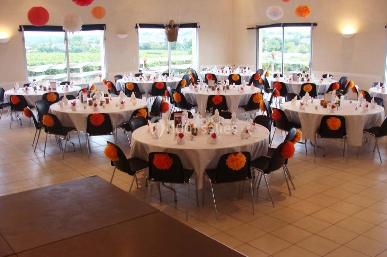 Salle décorée pour un événement, avec des tables rondes dressées et ornées de fleurs en papier colorées.