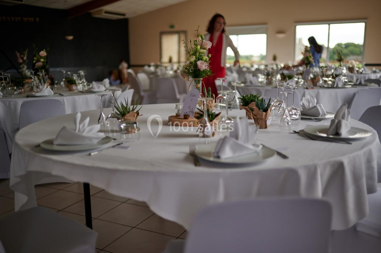 Salle de réception avec tables rondes dressées, nappes blanches, décorations florales et lumière naturelle.