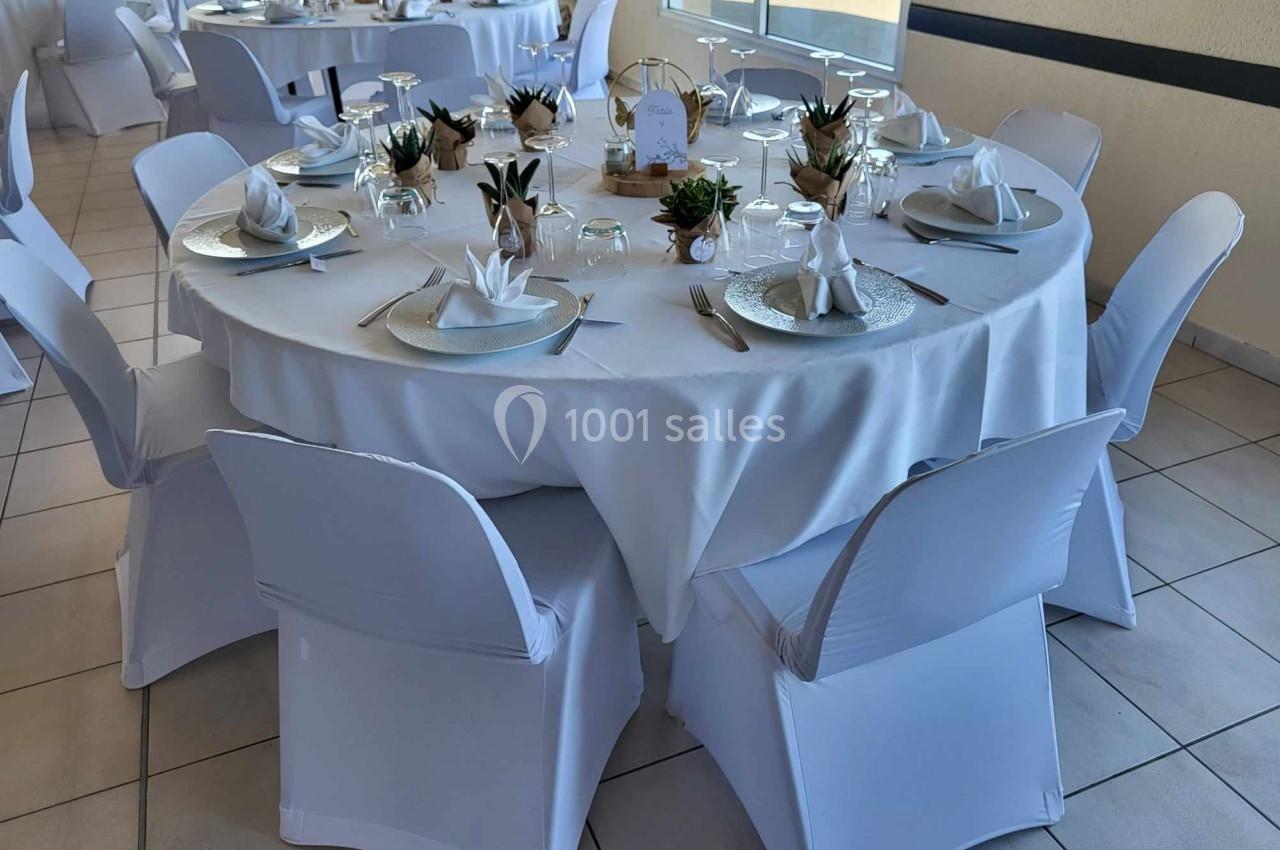 Table ronde décorée pour un événement, avec nappes blanches, serviettes pliées, vaisselle élégante et petites plantes.