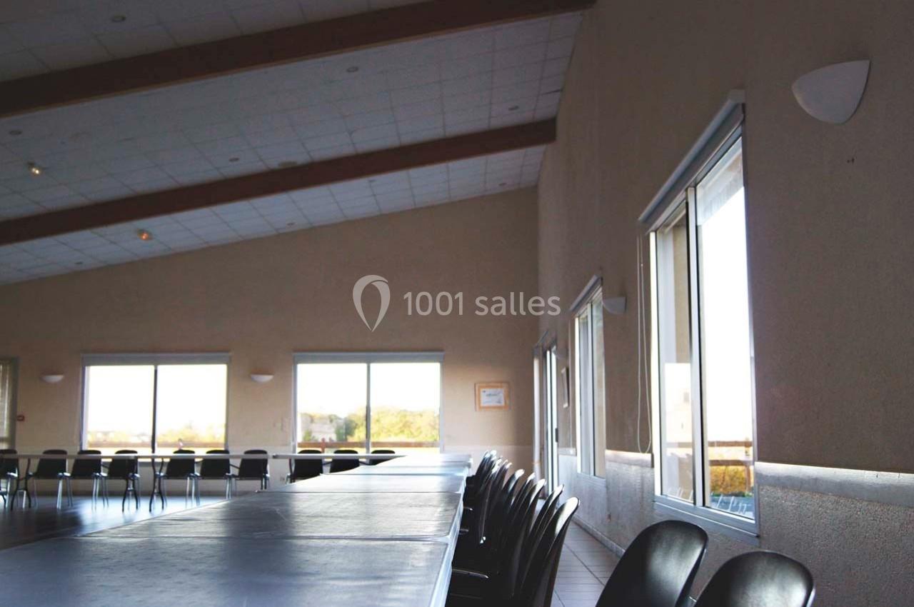 Salle lumineuse avec de grandes fenêtres, des tables alignées et des chaises noires disposées autour.
