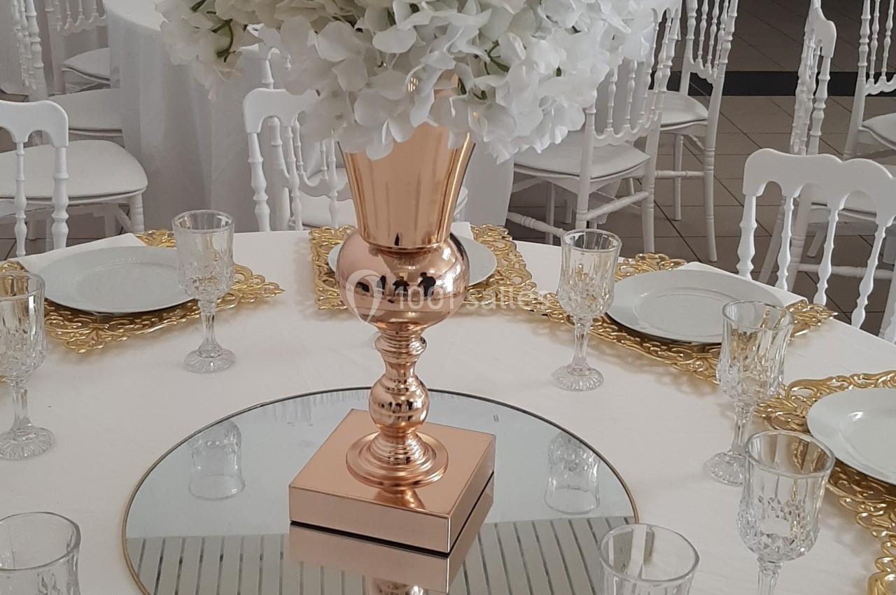 Centre de table avec vase doré, bouquet de fleurs blanches et roses, entouré de vaisselle élégante sur une nappe blanche.