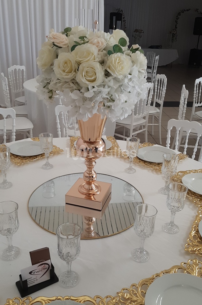 Centre de table avec vase doré, bouquet de fleurs blanches et roses, entouré de vaisselle élégante sur une nappe blanche.