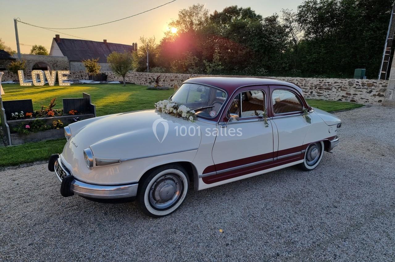 Voiture ancienne blanche et bordeaux décorée de fleurs, stationnée sur une allée gravillonnée au coucher du soleil.