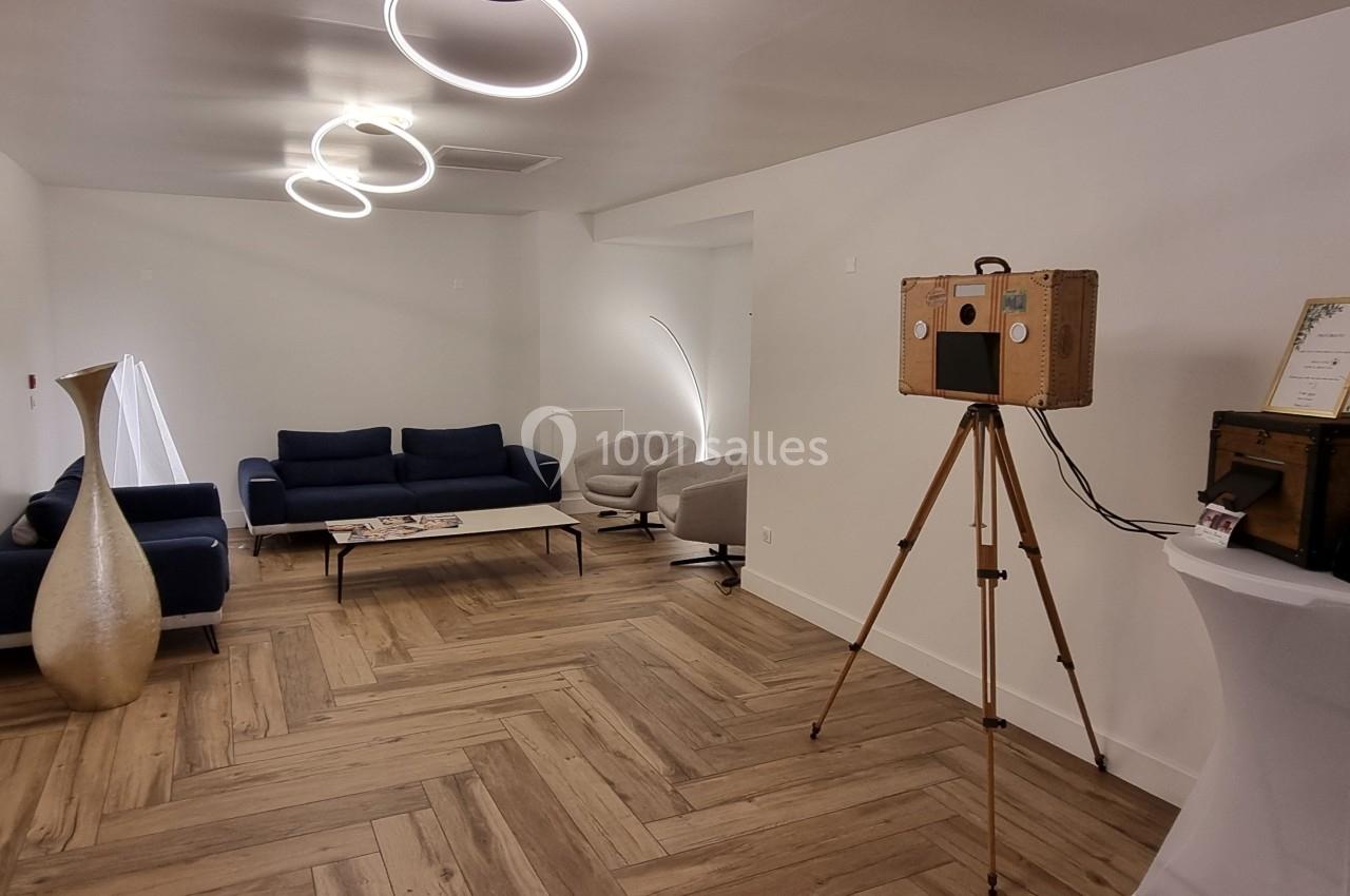 Salle lumineuse avec parquet en bois, canapé bleu, fauteuils clairs, vase décoratif et borne photo vintage sur trépied.