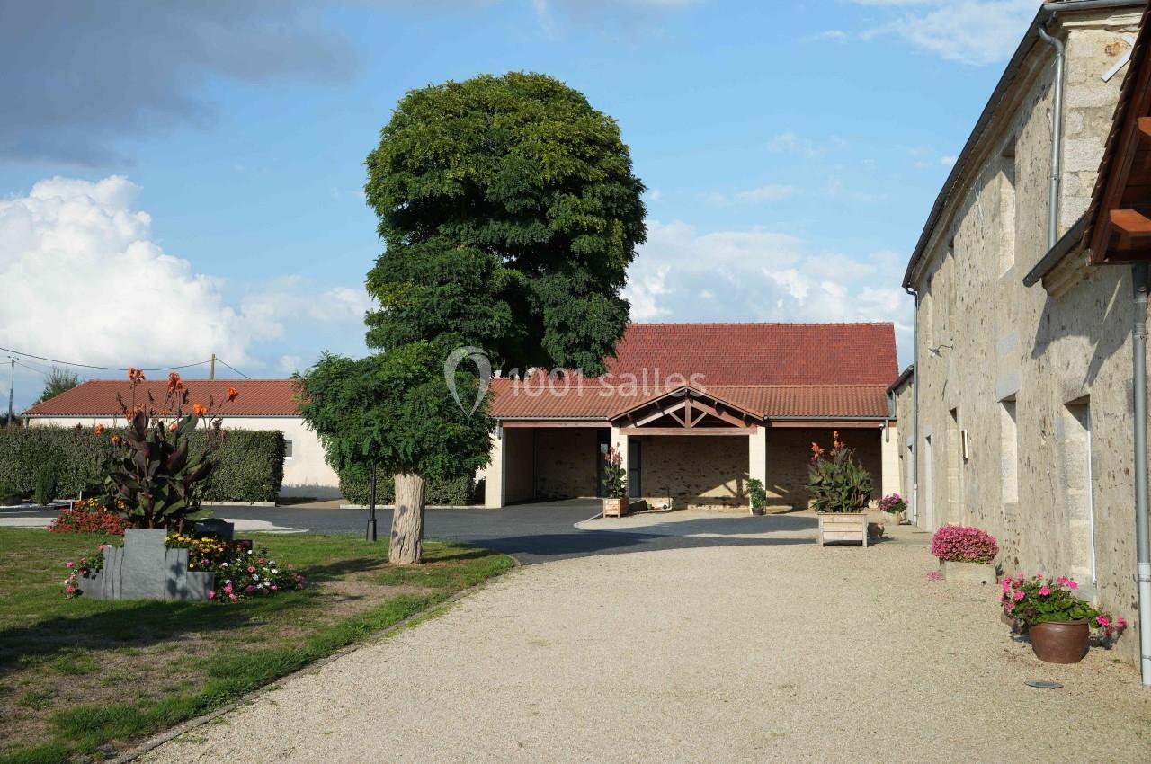 Cour d'une propriété rurale avec bâtiments en pierre, toit rouge, arbre central et fleurs décoratives.