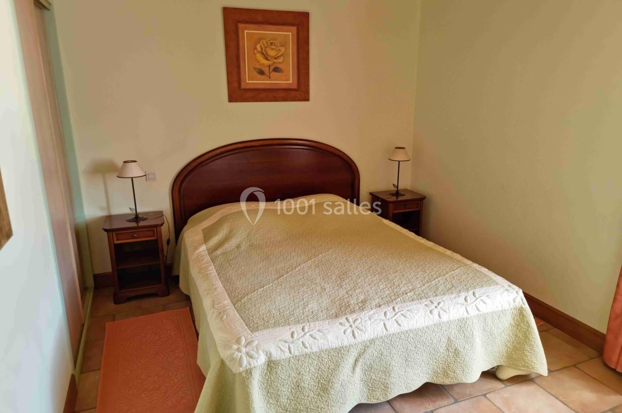 Lit double avec couvre-lit beige dans une chambre lumineuse, deux tables de chevet avec lampes et un tableau au mur.