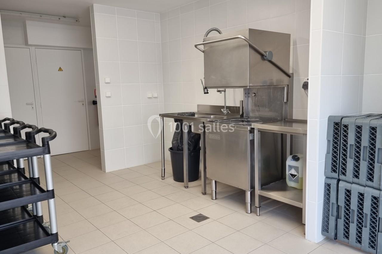 Cuisine professionnelle avec évier en inox, lave-vaisselle industriel, chariots noirs et casiers empilés sur sol carrelé.