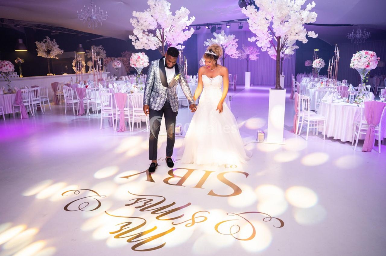Un couple de mariés marche main dans la main sur une piste de danse décorée, entourée de tables et d'arbres fleuris.