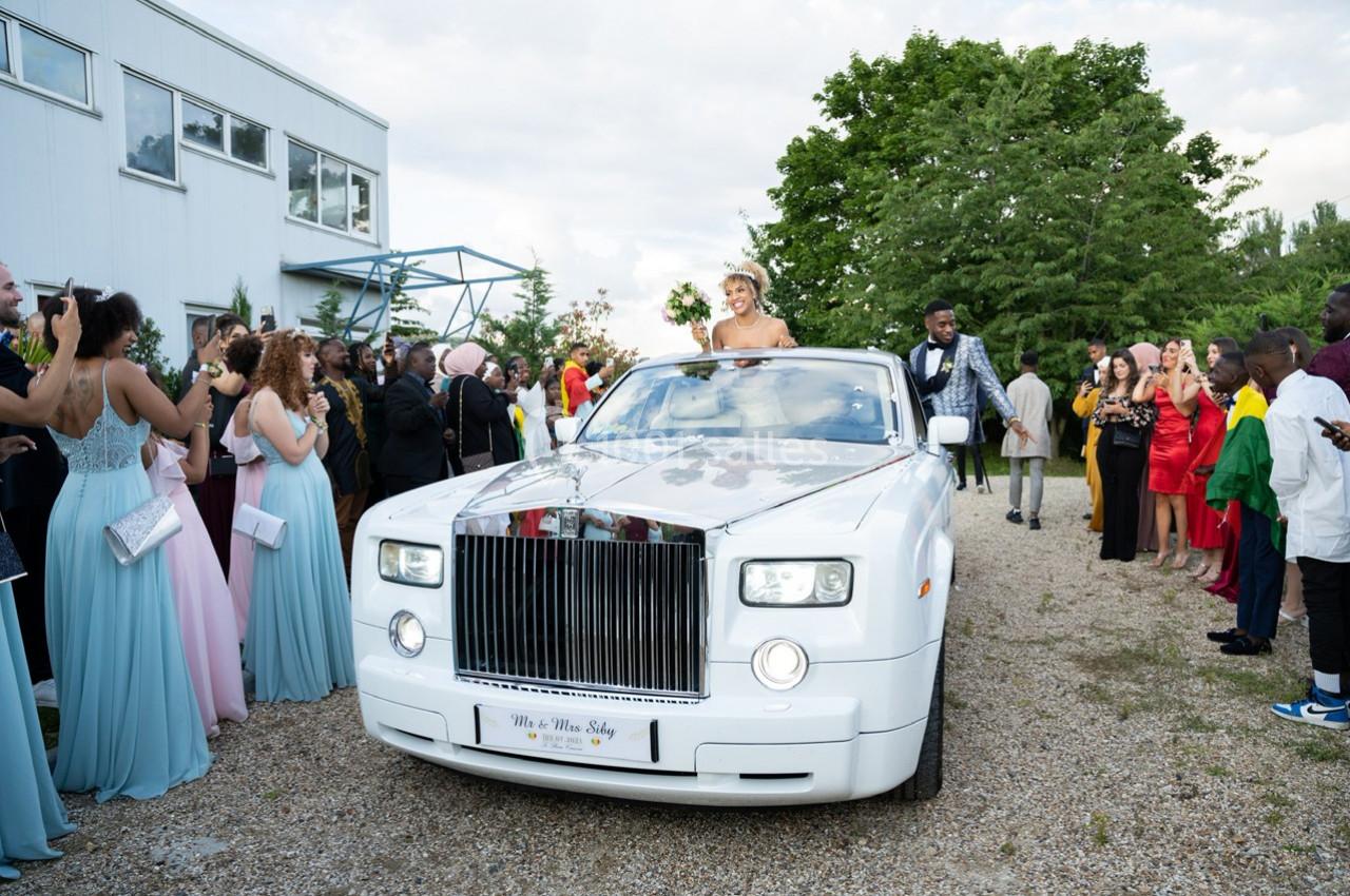 Une mariée et un marié dans une voiture blanche entourés d'invités en tenue de cérémonie.