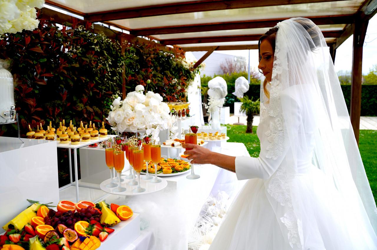 Une mariée en robe blanche se sert à un buffet décoré de fleurs blanches et de fruits sous une pergola.