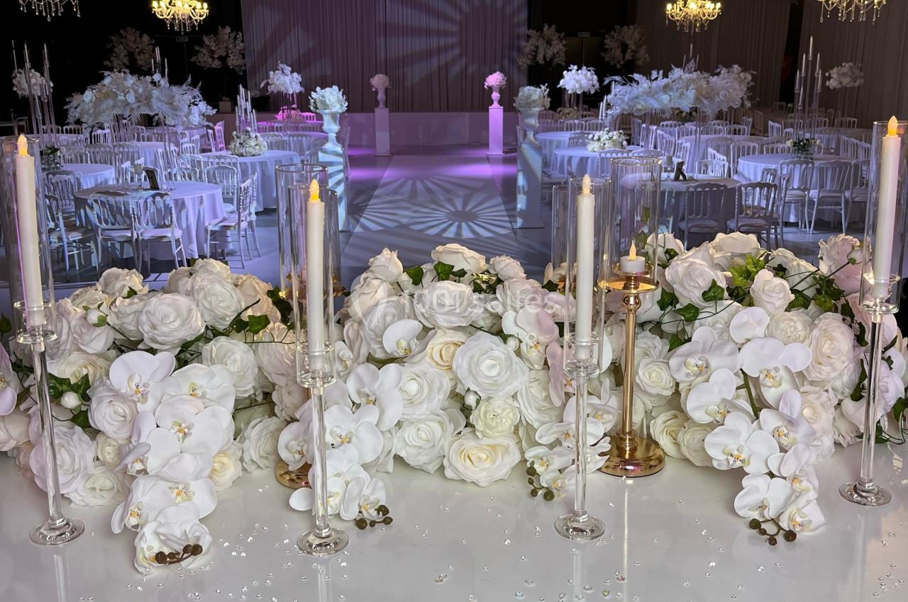 Salle de réception élégante décorée de fleurs blanches, chandeliers et bougies, avec une piste de danse éclairée.