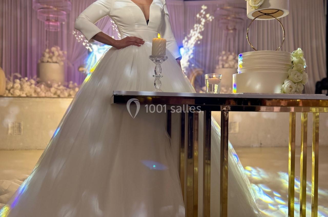 Une femme en robe de mariée blanche se tient près d'une table avec un gâteau décoré de fleurs blanches.
