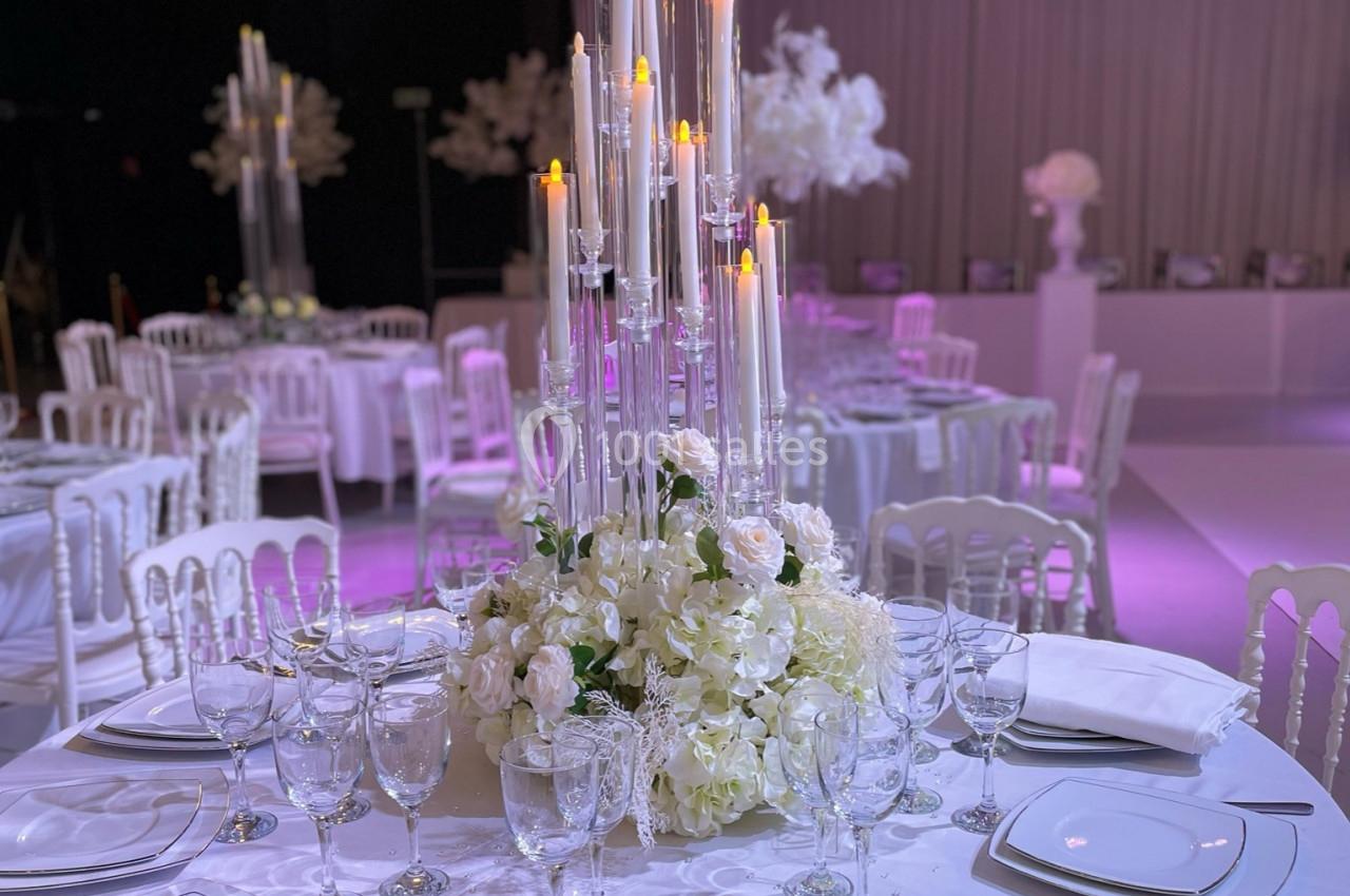 Centre de table avec chandeliers et fleurs blanches, entouré de tables dressées dans une salle de réception élégante.